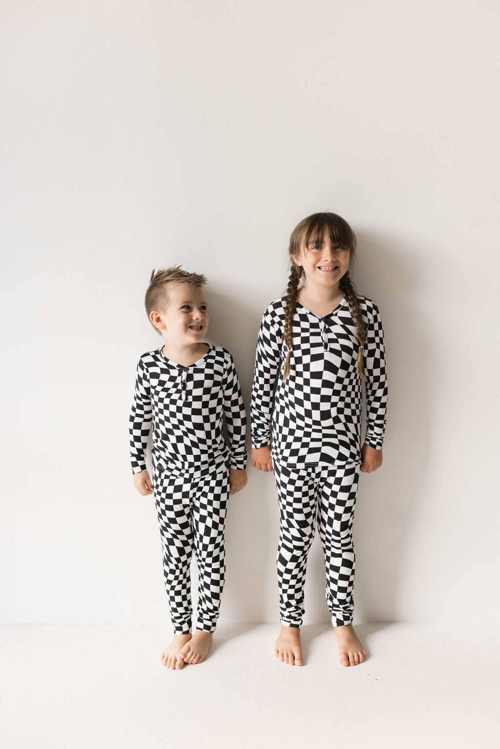 Two children are standing against a plain white wall, both smiling and barefoot. They are wearing matching Bamboo Two Piece Pajamas in Black & White Wavy Checkerboard by forever french baby. The child on the left has short hair, while the child on the right has long hair tied in two braids.