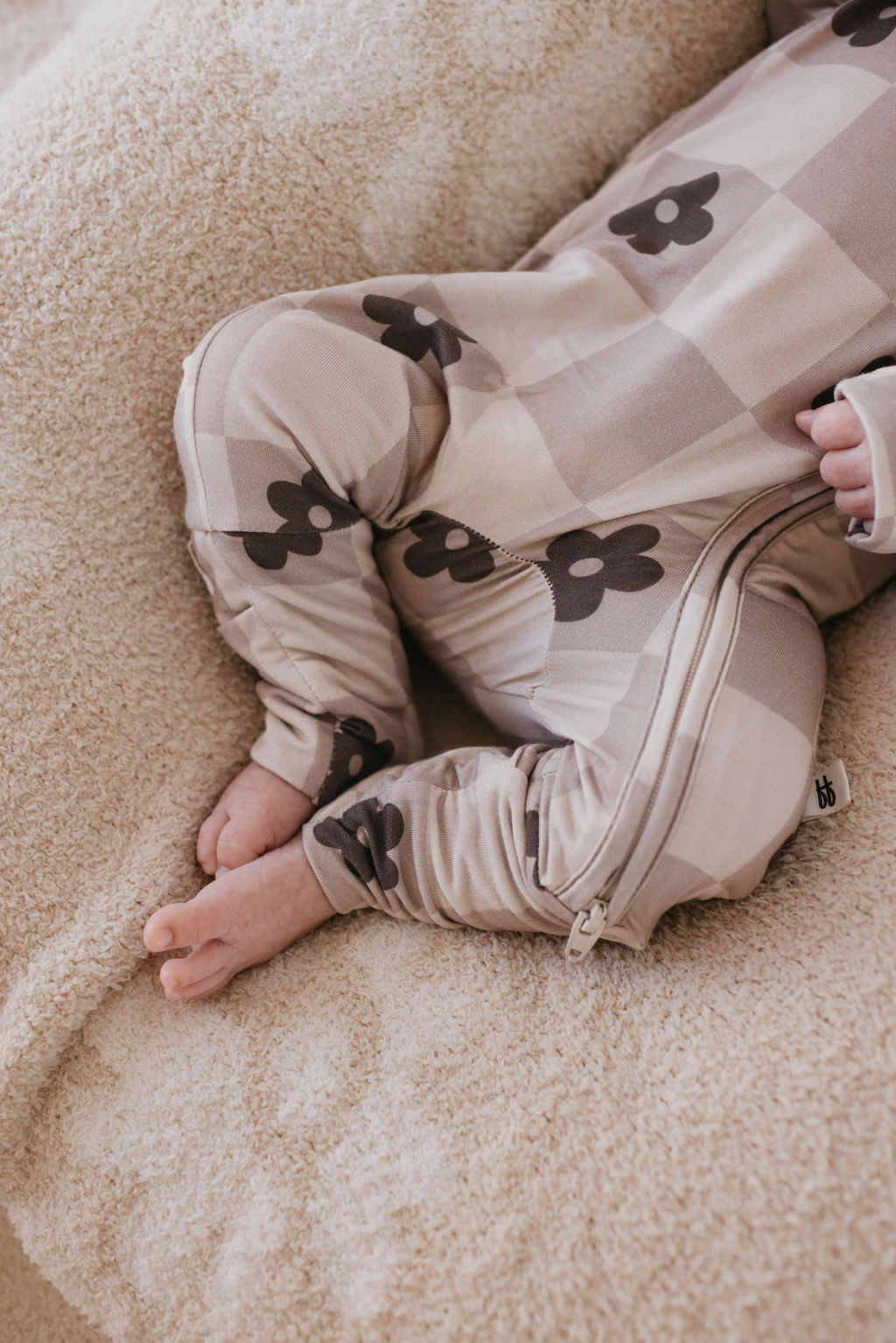 A baby lies on a soft, beige blanket, dressed in breathable sleepwear adorned with floral patterns. Only the feet and lower body are visible, drawing attention to the legs. The Bamboo Zip Pajamas from forever french baby feature a zipper running along the side, offering a blend of comfort and style.