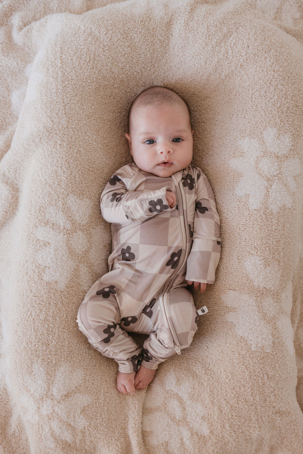 A baby lies on a soft, patterned blanket, wearing Forever French Baby's breathable Bamboo Zip Pajamas in the Flower Child design—a beige and brown checkered onesie adorned with floral patterns. The baby gazes up with one arm bent and the other resting by their side.