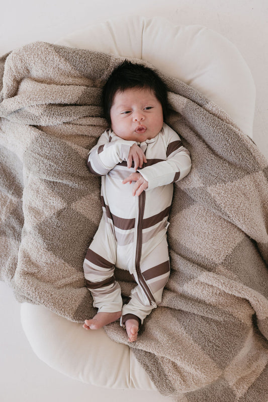 A baby wearing the Woodland Stripes Bamboo Zip Pajamas from forever french baby lies on a soft, beige, textured blanket. The baby looks up with their arms crossed over their chest, resting comfortably on the cozy surface.