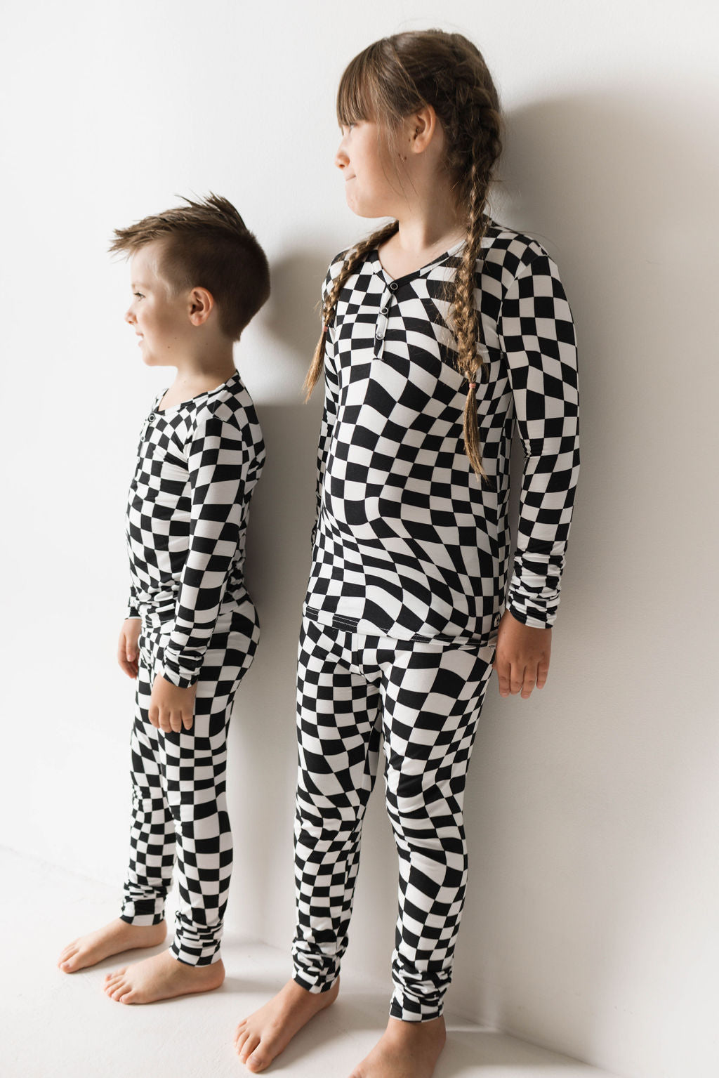 Two children stand against a white wall, wearing matching forever french baby Bamboo Two Piece Pajamas in Black & White Wavy Checkerboard pattern. The taller child has long braided hair, while the shorter child has short hair. Both look to the right, smiling slightly.