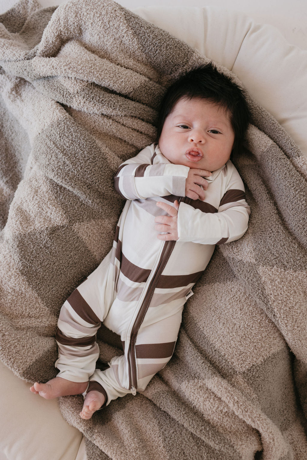 A baby lies on a beige blanket, clad in hypo-allergenic pajamas from forever french baby—a Bamboo Zip Pajamas featuring a white onesie with Woodland Stripes. The little one, with dark hair, gazes upward curiously, one hand gently touching their face.