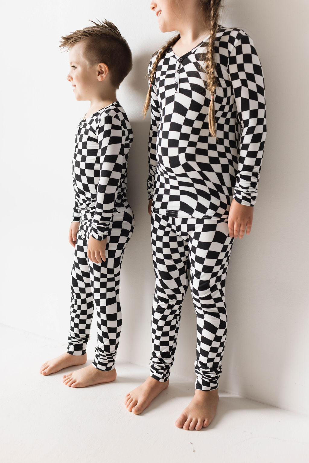 Two children stand against a white wall, wearing forever french baby's Bamboo Two Piece Pajamas in Black & White Wavy Checkerboard. The child on the left has short hair styled upwards, while the child on the right has long hair braided into two braids. Both are barefoot and smiling, enjoying their soft, hypo-allergenic clothing.