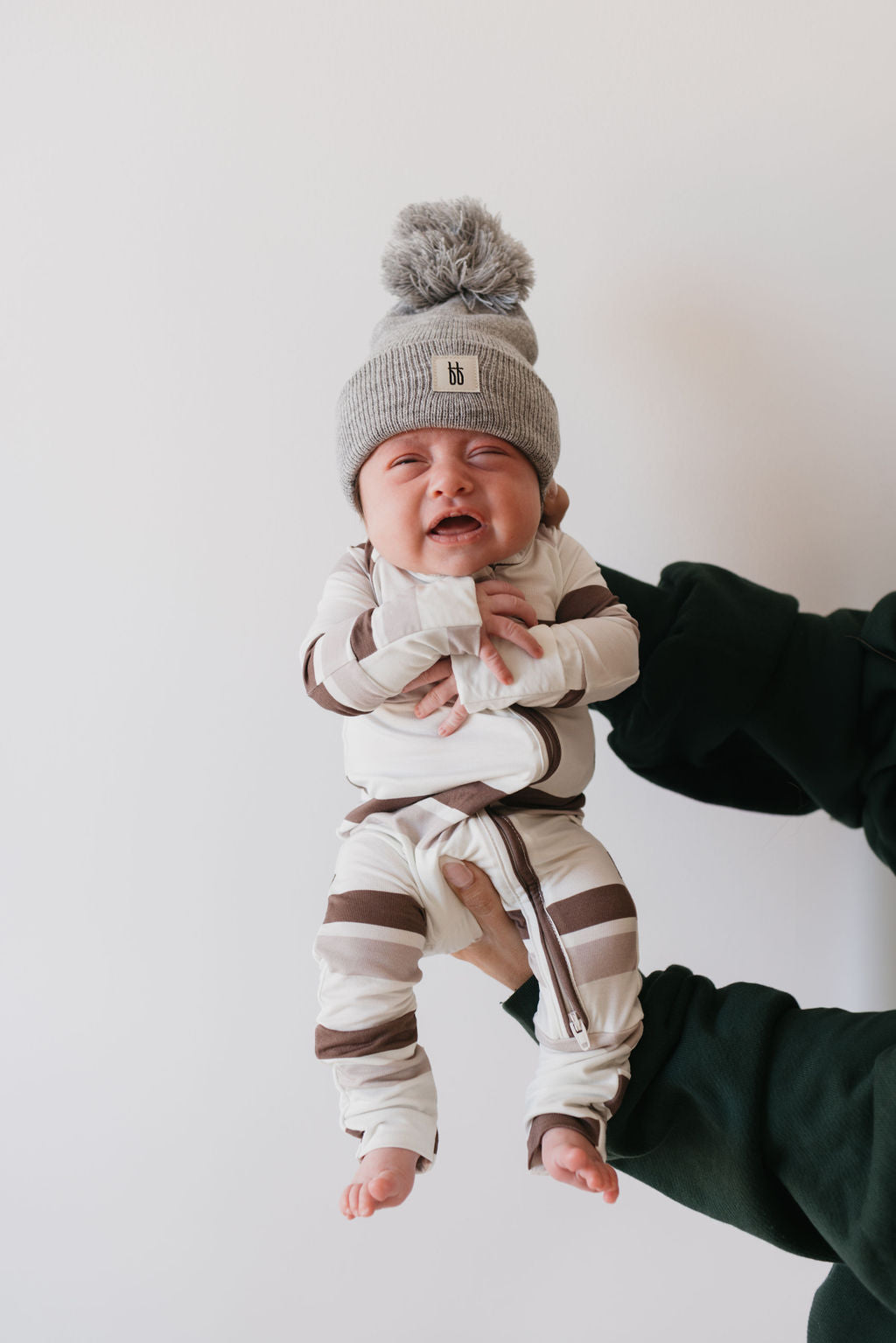 A baby dressed in forever french baby's Bamboo Zip Pajamas in Woodland Stripes, complemented by a gray pom-pom hat, is held by someone wearing a dark green sweater. With a joyful expression, the baby stands out against the plain white background.