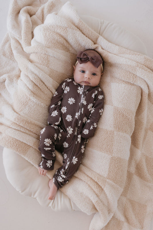 A baby is dressed in the Midnight Meadow bamboo zip pajamas from Forever French Baby, paired with a matching headband, and lies on a soft, beige checked blanket.