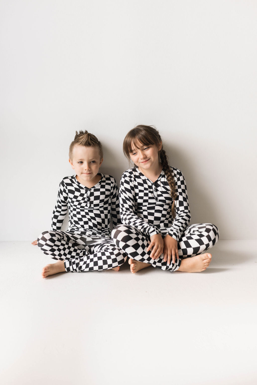 Two children sit side by side against a white wall, both dressed in matching Bamboo Two Piece Pajamas in a Black & White Wavy Checkerboard pattern from forever french baby. The child on the left has short hair styled into a small mohawk, while the child on the right has long hair with bangs.