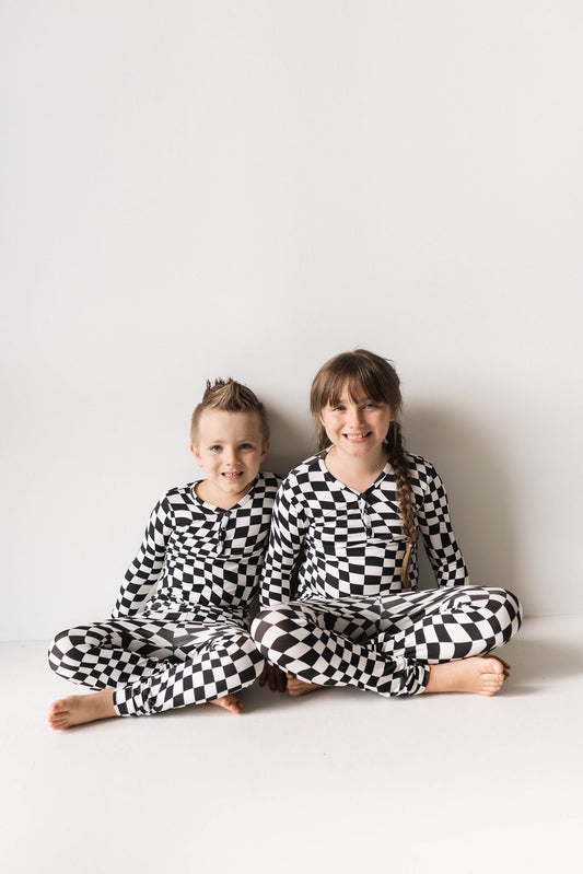 Two children sit side by side on the floor against a plain white background. They are wearing matching Bamboo Two Piece Pajamas in a Black & White Wavy Checkerboard pattern from forever french baby, made from breathable fabric. The child on the left has short hair, while the child on the right has long hair tied into two braids. Both are smiling.