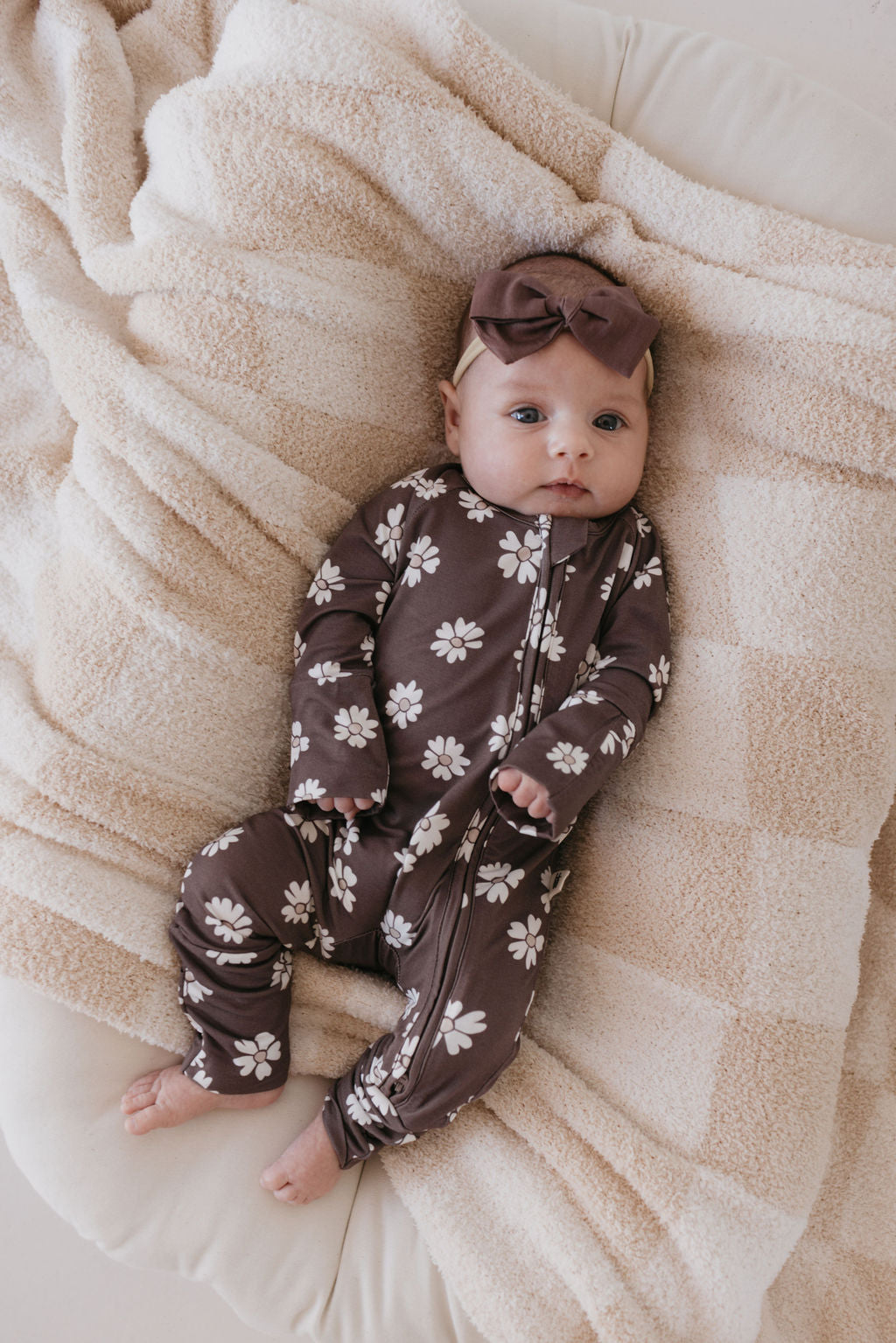 A baby dressed in a Midnight Meadow Bamboo Zip Pajamas by forever french baby, featuring a brown floral design made from breathable fabric, with a matching headband, lies comfortably on a soft beige blanket.