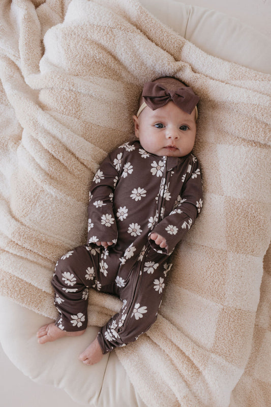 A baby dressed in a Midnight Meadow Bamboo Zip Pajamas by forever french baby, featuring a brown floral design made from breathable fabric, with a matching headband, lies comfortably on a soft beige blanket.