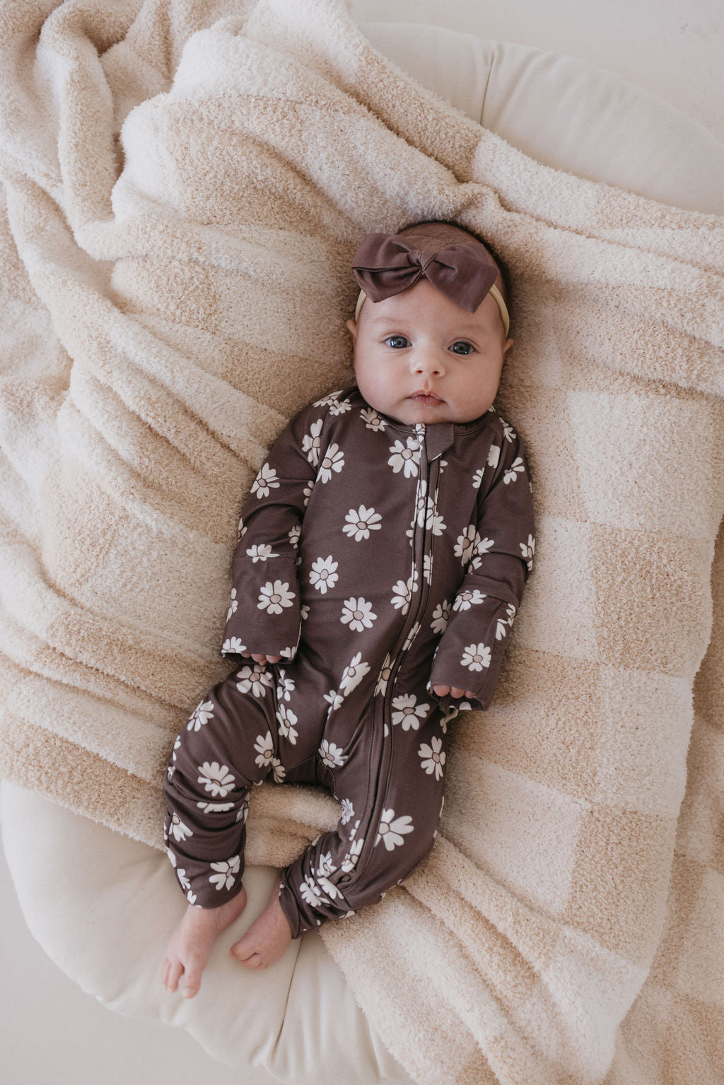 A baby lies on a soft, beige checkered blanket, wearing a stylish brown Bamboo Zip Pajamas from the Midnight Meadow collection by forever french baby, featuring delicate white flower patterns and crafted from breathable fabric. A matching brown headband with a bow completes the look as the baby gazes up with a calm expression.