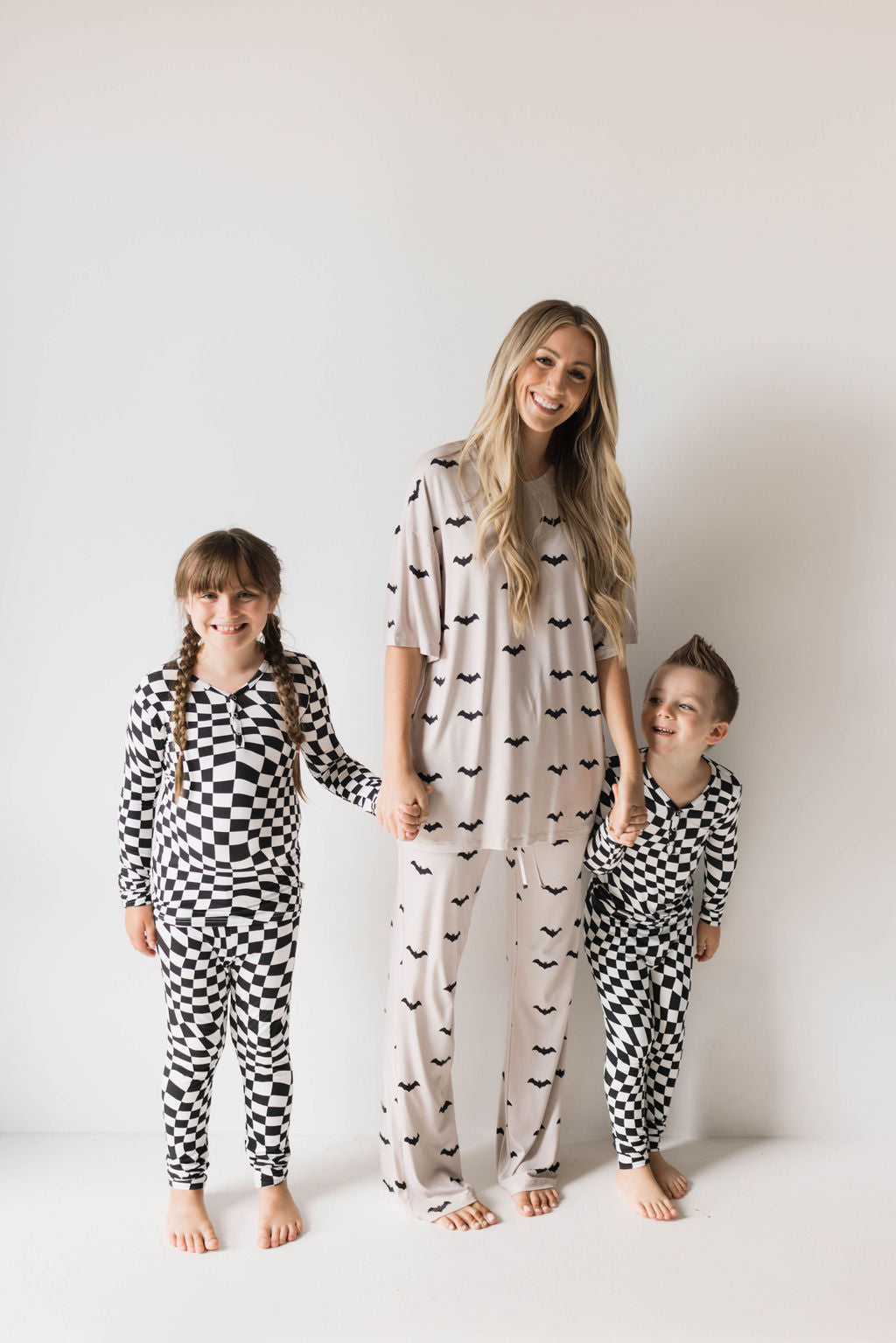 A woman stands barefoot in oversized "Short Sleeve Women's Bamboo Pajamas | It's Bats!" from forever french baby. She holds hands with two children on either side. The child on her left is dressed in hypo-allergenic black and white checkered pajamas, while the child on her right matches in a similar onesie.