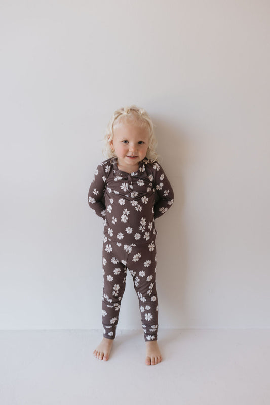 A small child with curly blonde hair stands barefoot against a white wall, wearing the Midnight Meadow Bamboo Two Piece Pajamas by forever french baby. The outfit's comfy and breathable fabric complements the child's cheerful expression as their hands are playfully hidden behind their back.