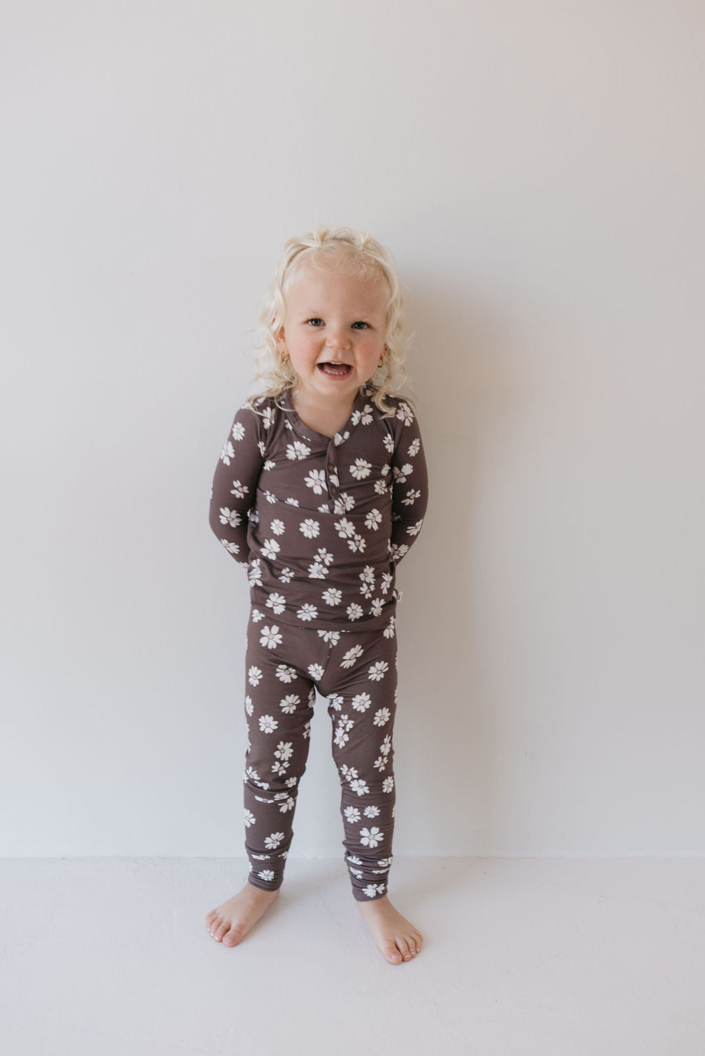 A young child with curly blonde hair stands against a white background, smiling while wearing the Midnight Meadow Bamboo Two Piece Pajamas from forever french baby. These hypo-allergenic pajamas, adorned with charming white daisy patterns on a brown fabric, are crafted from breathable bamboo material to ensure comfort and style for a restful night's sleep.