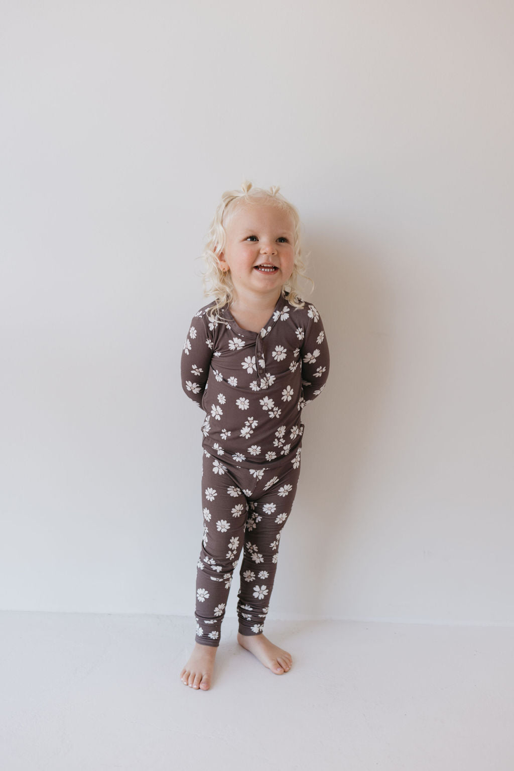 A smiling toddler with curly blonde hair wears the "Midnight Meadow" bamboo two-piece pajamas by Forever French Baby. The brown pajama set, adorned with white floral patterns, is made from hypo-allergenic and breathable bamboo fabric, offering comfort and ease as the child stands barefoot against a plain white background, looking upward.