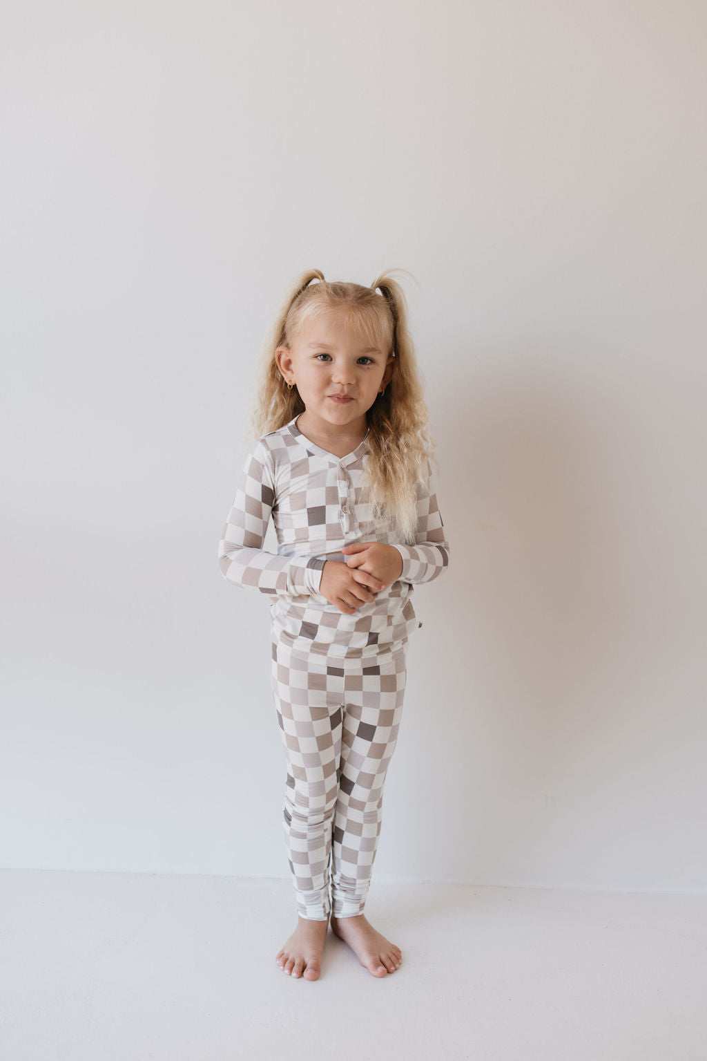 A young child with long blond hair stands barefoot in front of a plain white wall, wearing the Bamboo Two Piece Pajamas from forever french baby. The breathable set, titled "Into the Woods Checkerboard," features a matching checkered long-sleeve top and pants. They have a neutral expression with their hands clasped in front.