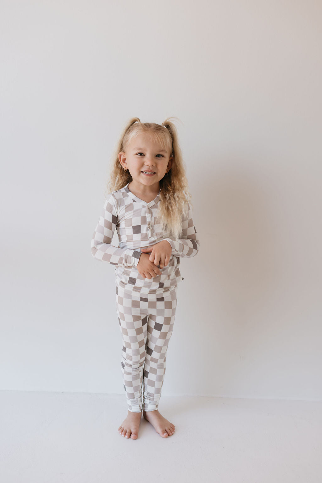 A young child with long blonde hair stands smiling against a plain backdrop, wearing the Bamboo Two Piece Pajamas | Into the Woods Checkerboard by forever french baby. These breathable pajamas are crafted from hypo-allergenic fabric and feature a beige and white checkerboard pattern. She stands barefoot, arms gently clasped in front.