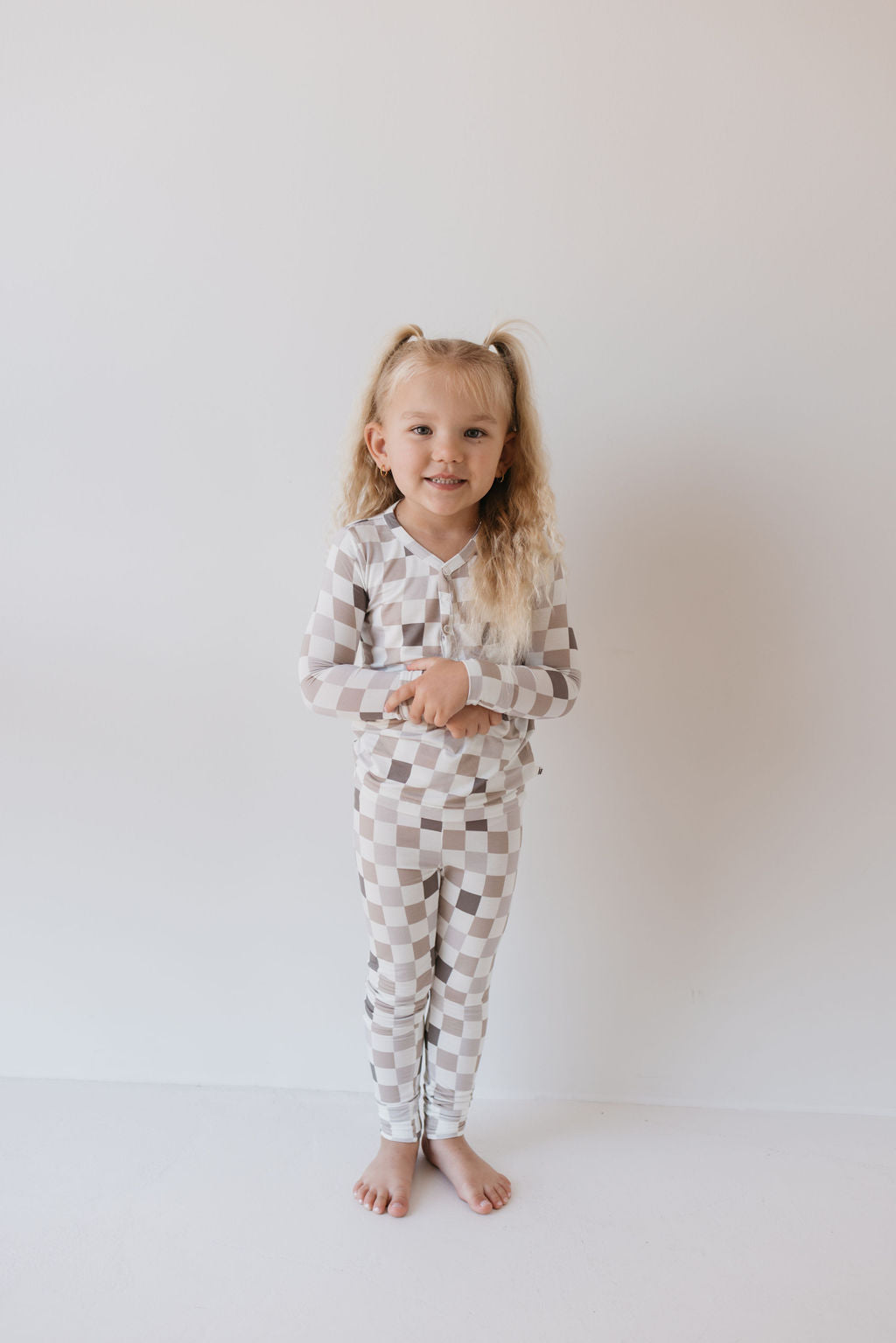 A young girl with long blonde hair stands barefoot against a plain white background, wearing the Bamboo Two Piece Pajamas by forever french baby. Her ensemble is the "Into the Woods Checkerboard" design, showcasing a matching outfit of long sleeves and pants in beige and white squares. Exhibiting a calm expression, she looks forward serenely.