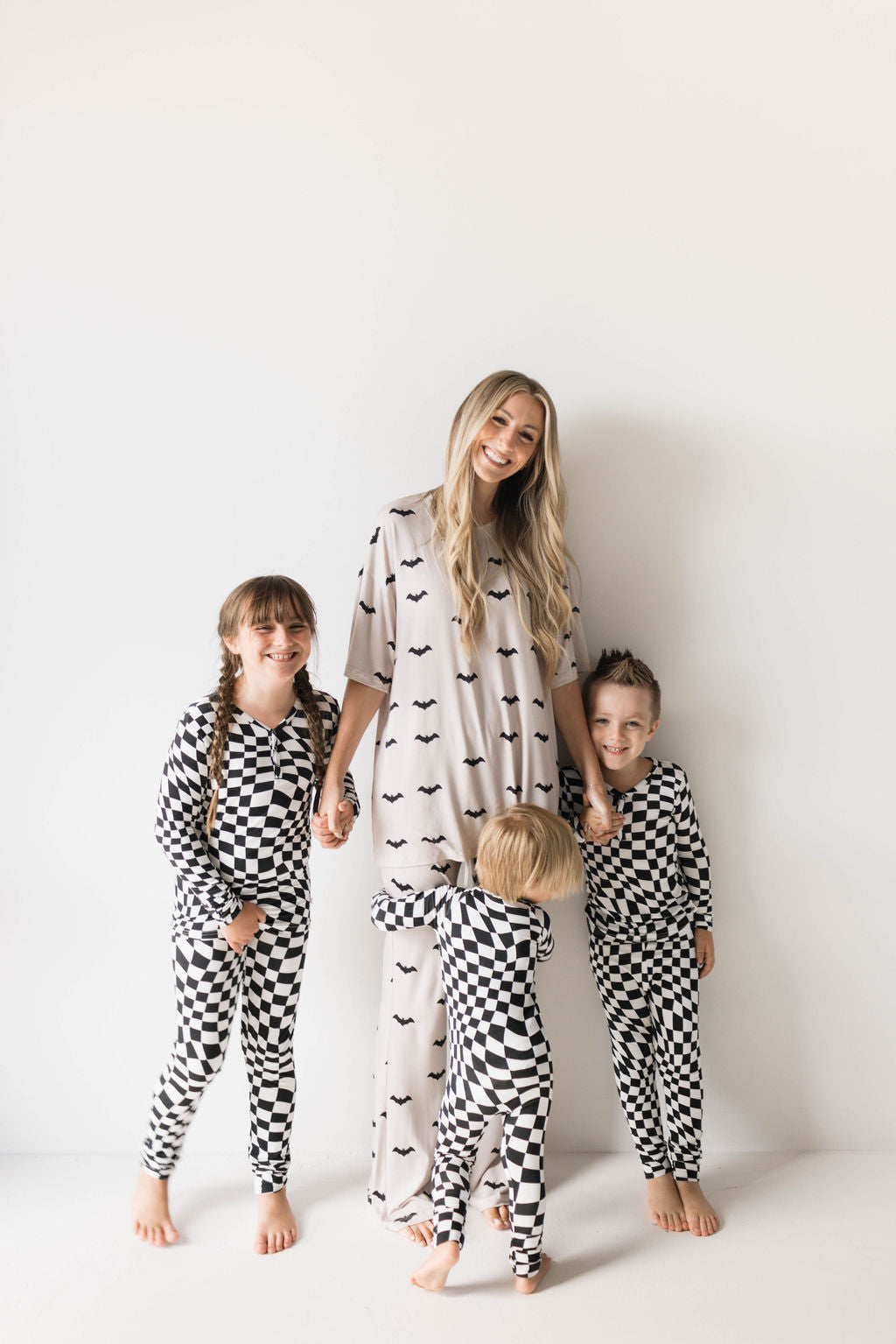 A woman with long blonde hair stands smiling, holding the hands of two children. She is wearing a light-colored, bat-themed outfit made from breathable fabric, and the children are dressed in Bamboo Two Piece Pajamas | Black & White Wavy Checkerboard by forever french baby. Another child stands in front of her, centering the group.