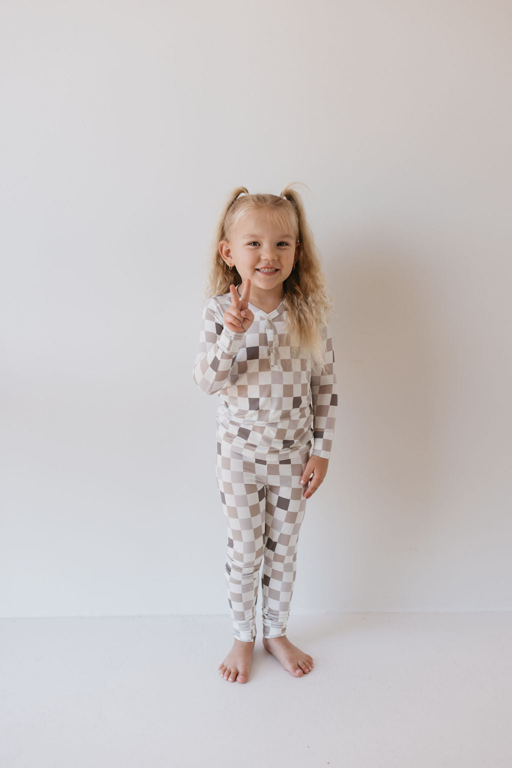 A young girl with long, curly blonde hair stands barefoot against a plain white background. She is wearing the Bamboo Two Piece Pajamas in "Into the Woods Checkerboard" from forever french baby, smiling as she holds up one finger.