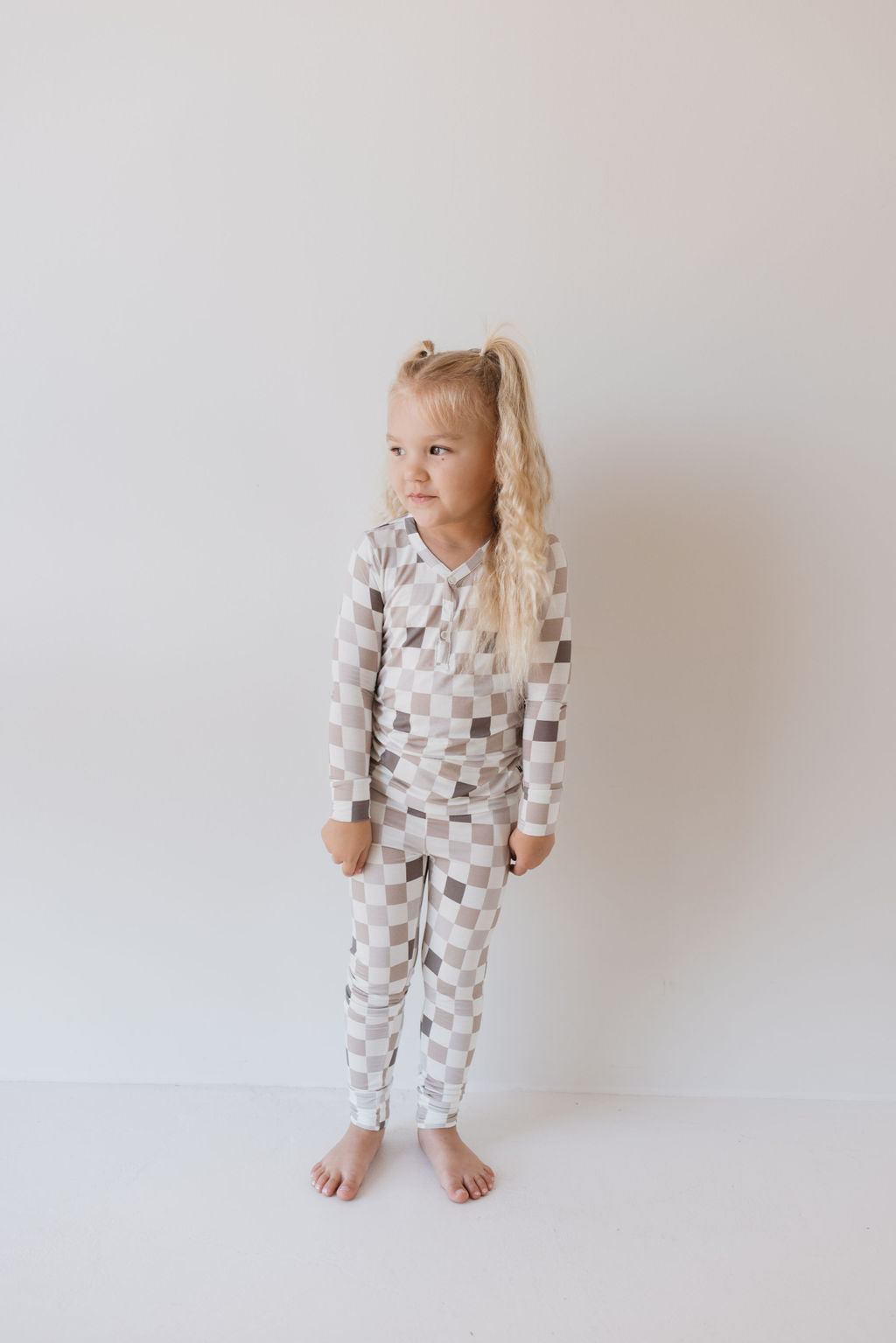 A young child with long blonde hair stands against a plain white background, wearing the Bamboo Two Piece Pajamas from forever french baby in a tasteful blend of beige and white. The Into the Woods Checkerboard design, made from hypo-allergenic bamboo fabric, perfectly complements their barefoot stance as they gaze off to the side with a neutral expression.