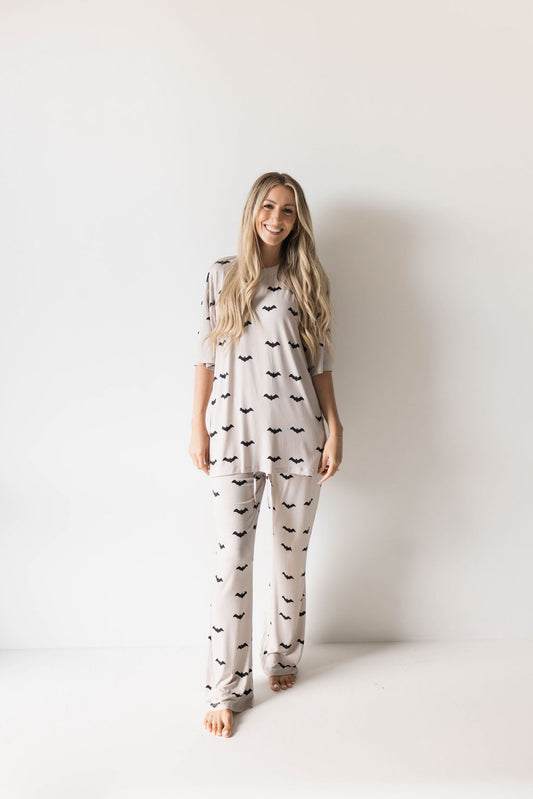 A smiling woman with long, blonde hair stands against a white background. She is wearing an oversized Short Sleeve Women's Bamboo Pajamas from forever french baby, featuring the "It's Bats!" pattern of small, black bats. The light-colored pajamas include a short-sleeve top and long pants. She is barefoot.