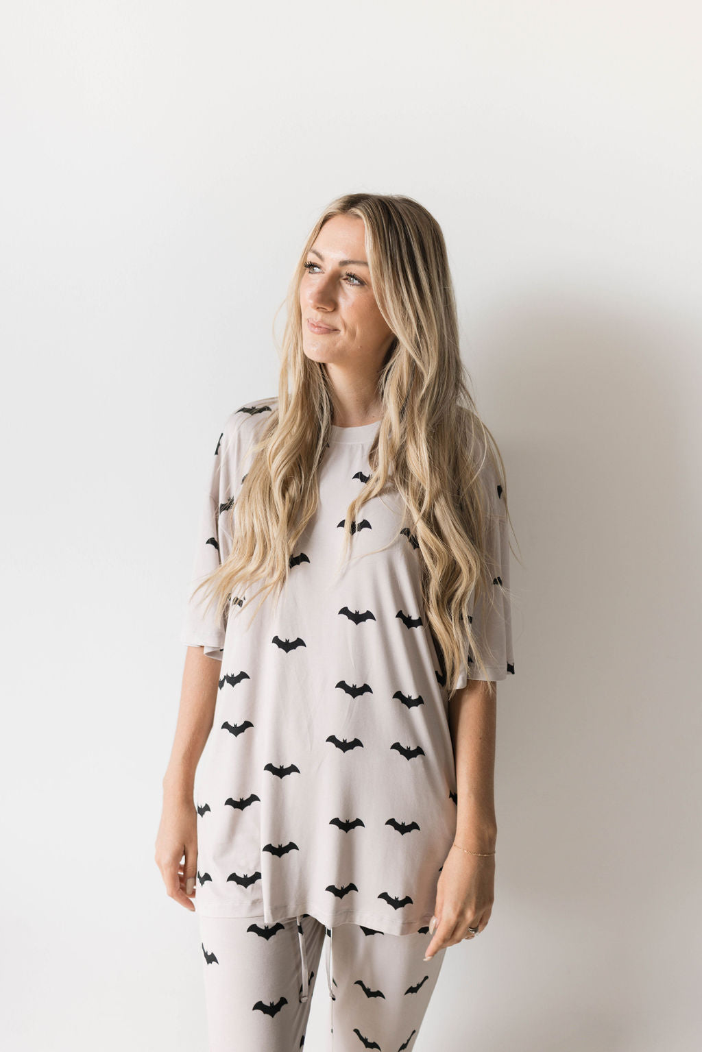 A person with long, blonde hair stands against a white background, dressed in an oversized "Short Sleeve Women's Bamboo Pajamas | It's Bats!" set from forever french baby. The light-colored ensemble features a black bat pattern and is hypo-allergenic, offering both style and comfort. They gaze to the side with a neutral expression.