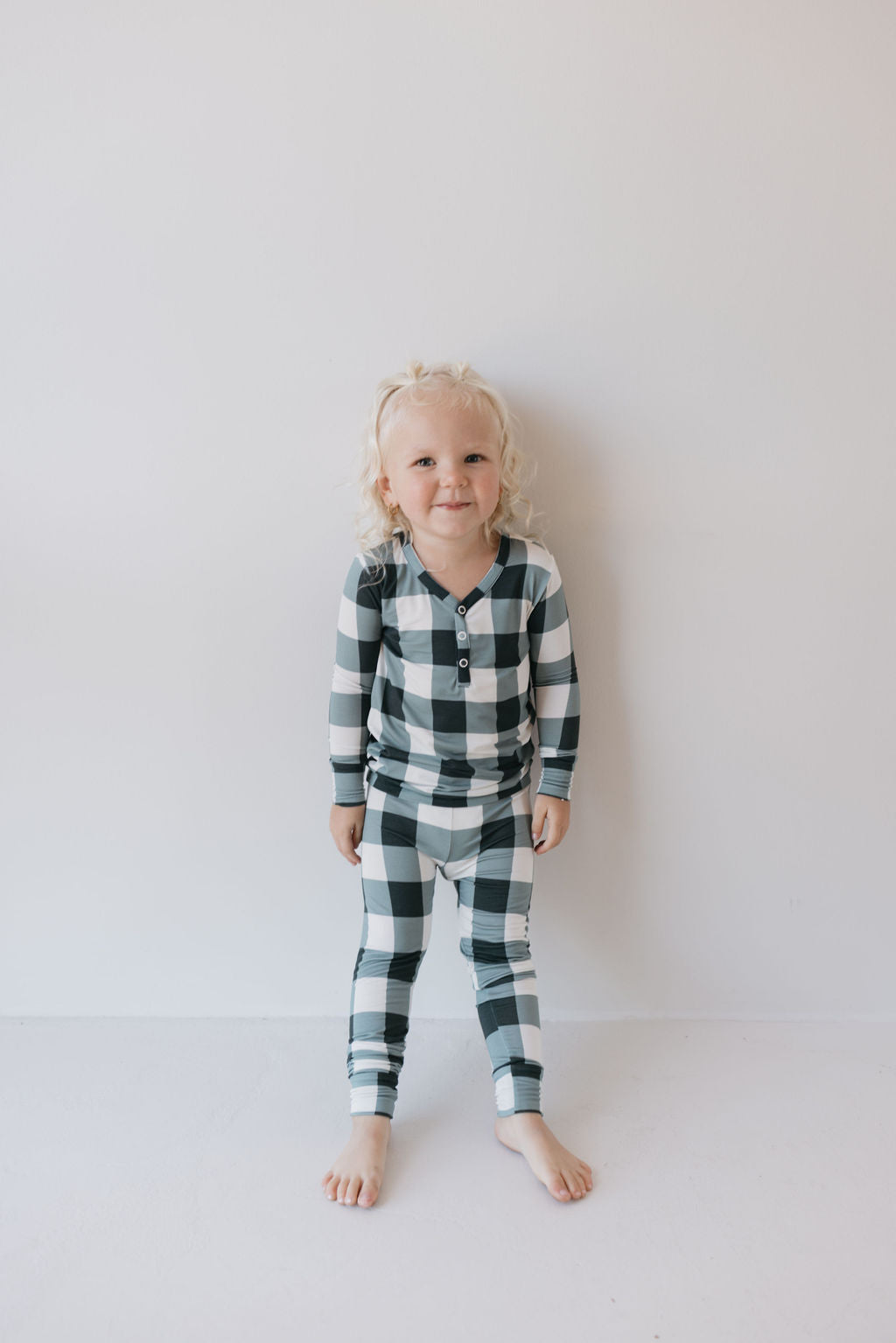 A young child with curly blond hair stands barefoot against a white background, wearing the forever french baby Bamboo Two Piece Pajamas in Evergreen Gingham. These pajamas offer hypo-allergenic comfort for a peaceful night's sleep with their breathable bamboo fabric.