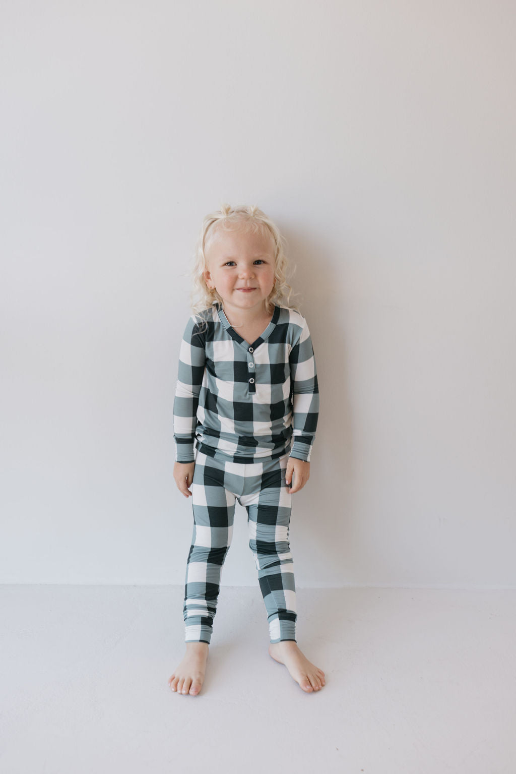 A small child with curly blonde hair stands against a plain white wall, dressed in the Bamboo Two Piece Pajamas in Evergreen Gingham by forever french baby. The outfit consists of a matching long-sleeve shirt and pants featuring a black and white checkered pattern made from breathable sleepwear fabric, and they are barefoot.