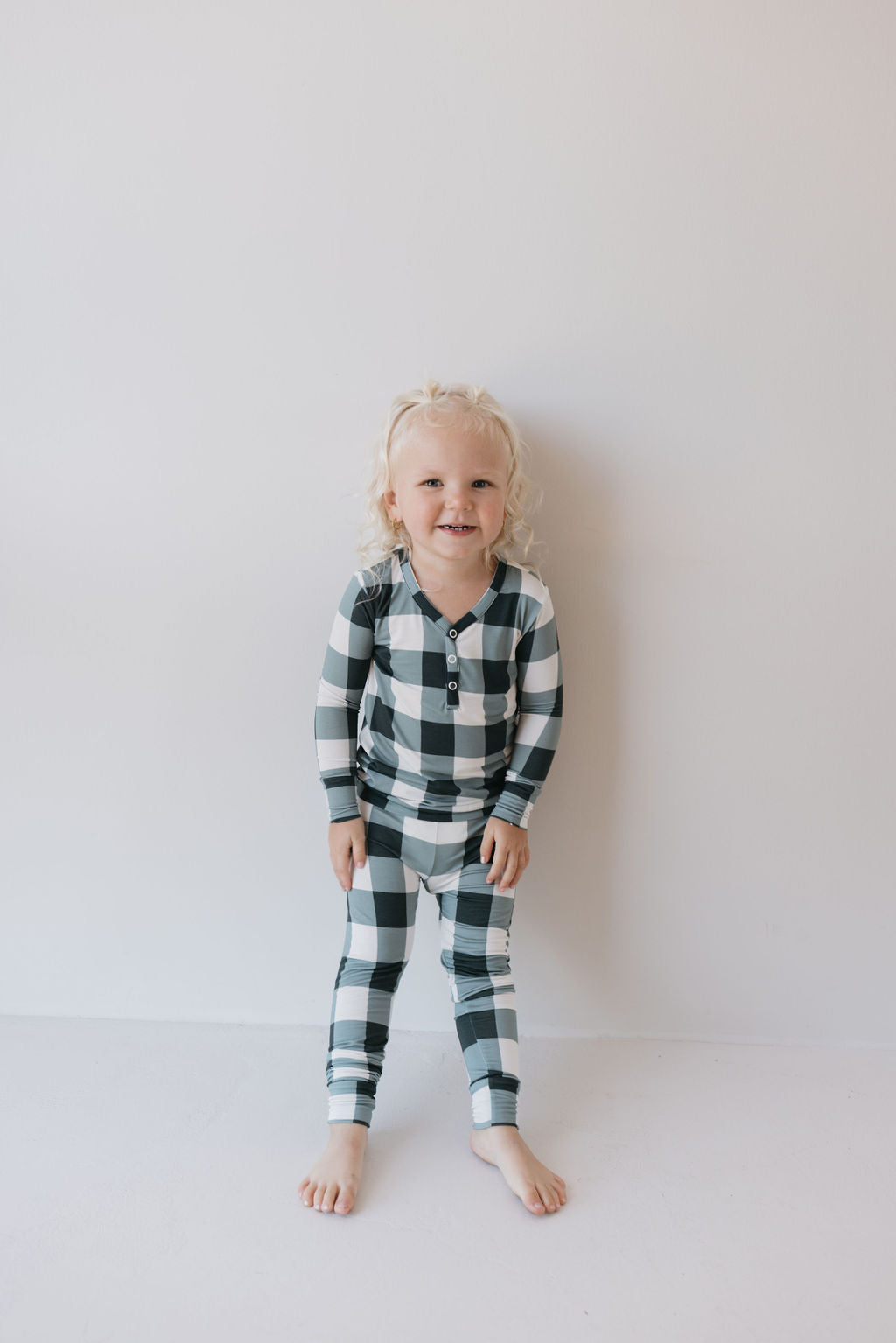 A small child with curly blonde hair is smiling, wearing the Evergreen Gingham Bamboo Two Piece Pajamas by forever french baby. They are barefoot and standing against a plain white background.