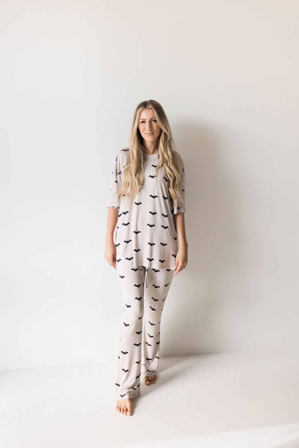 A woman with long, wavy hair stands barefoot against a plain white backdrop, smiling slightly. She is wearing an oversized pajama set consisting of a loose-fitting shirt and pants in light-colored fabric with a black abstract pattern from the Short Sleeve Women's Bamboo Pajamas | It's Bats! collection by forever french baby.