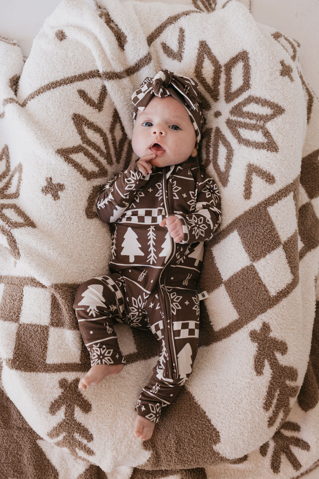 A baby dressed in a brown outfit with winter-themed patterns, such as trees and snowflakes, lies on a fluffy blanket. The child gazes upward with a hand near their mouth, wearing a Forever Fair Isle Bamboo Head Wrap by forever french baby—an adorable accessory for babies and toddlers that perfectly matches their cozy attire.