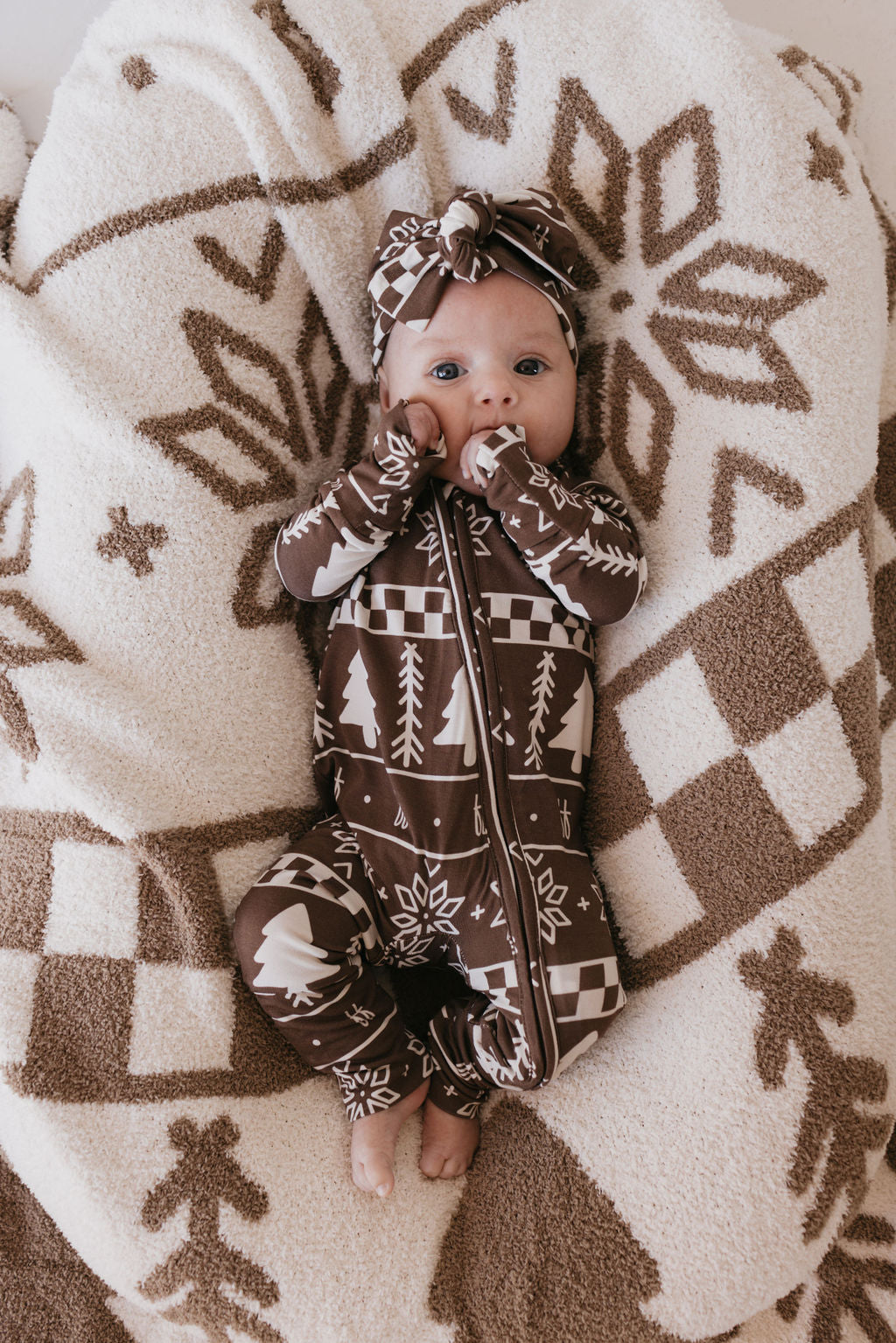 A baby lies on a large, patterned cushion, dressed in a brown and white jumpsuit with tree and snowflake designs. Sporting the Bamboo Head Wrap from forever french baby in the Forever Fair Isle pattern, the little one keeps their hands near their mouth amid cozy, winter-themed decor.