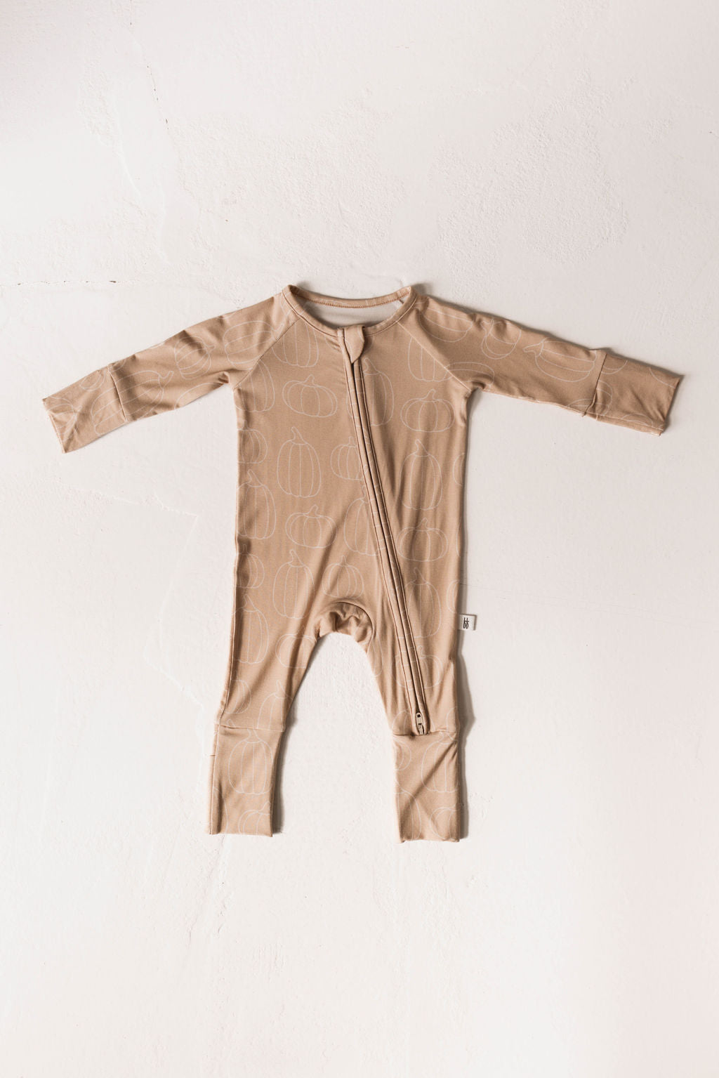 The Bamboo Zip Pajamas | Pumpkin Pie by forever french baby is a light brown baby onesie made from breathable, hypo-allergenic fabric. It features a subtle pumpkin pattern and long sleeves, with a zipper running from the neck down to the left leg. The onesie is displayed flat on a white background.