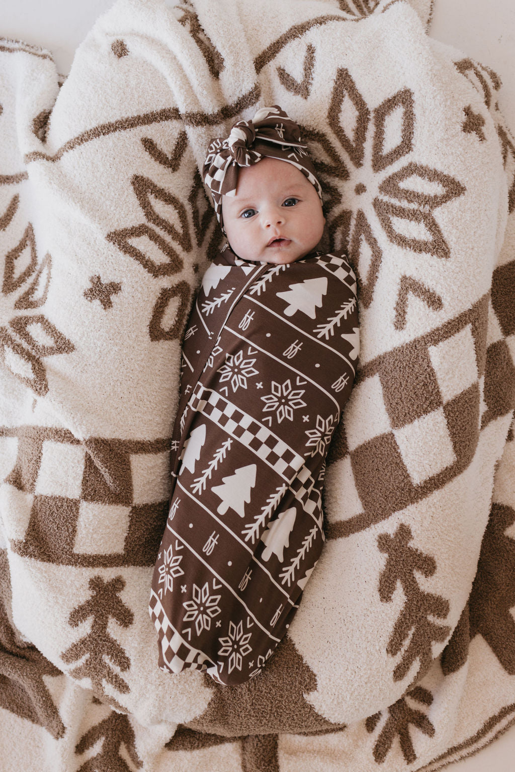 A baby lies wrapped in a breathable, hypo-allergenic Forever Fair Isle bamboo swaddle by forever french baby, featuring brown and white tree and geometric designs on a fluffy matching blanket. The baby wears a coordinating headband and gazes up, enveloped in cozy, winter-themed fabric.