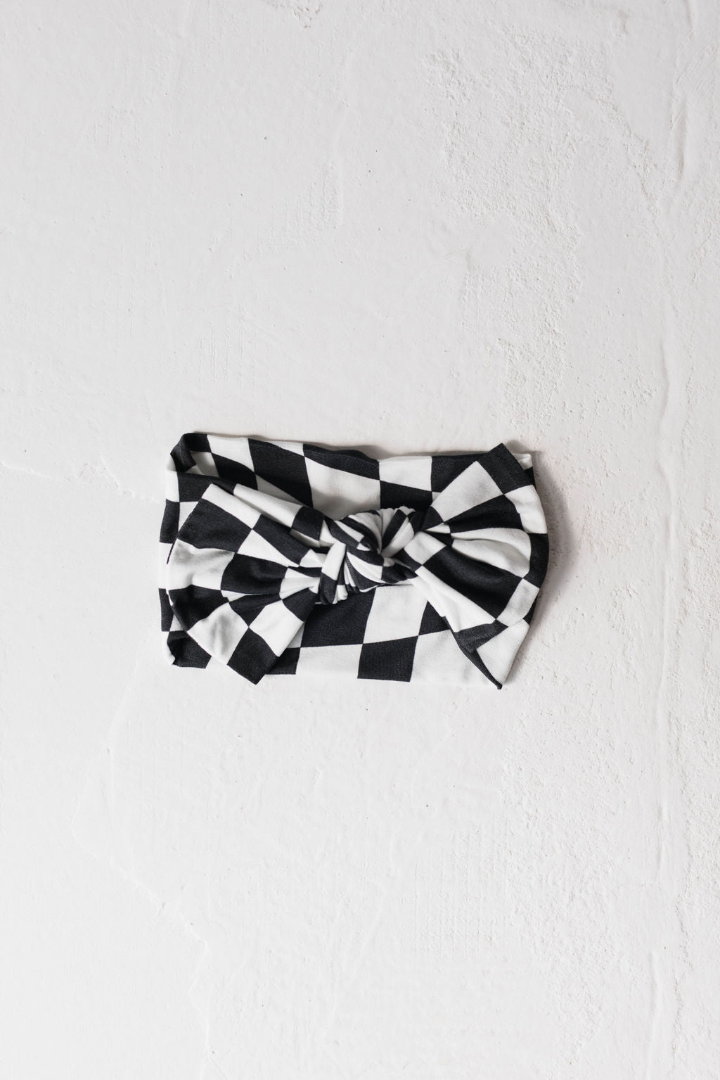 A black and white wavy checkerboard headband, perfect for babies and toddlers, is laid flat on a textured white surface. The headband, known as the Bamboo Head Wrap by forever french baby, features a charming knotted design at the center.