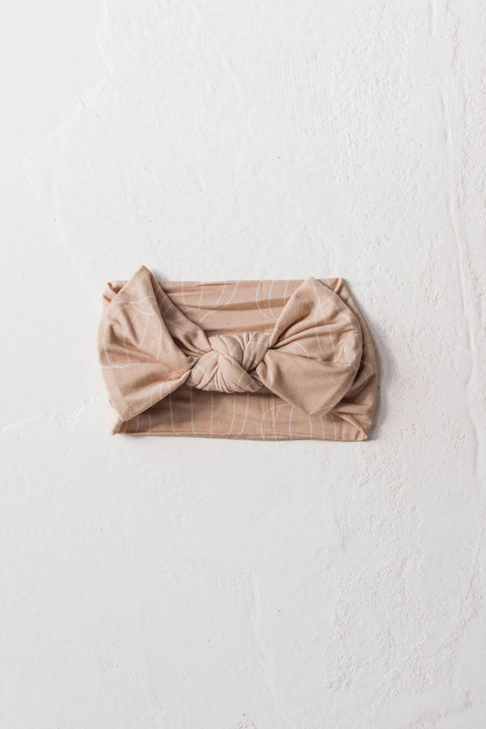 A Pumpkin Pie-colored, knotted headband featuring subtle white lines forming a leaf-like pattern lies flat on a textured, off-white background. This chic bamboo head wrap from forever french baby is wrapped elegantly and showcases a central knot design.