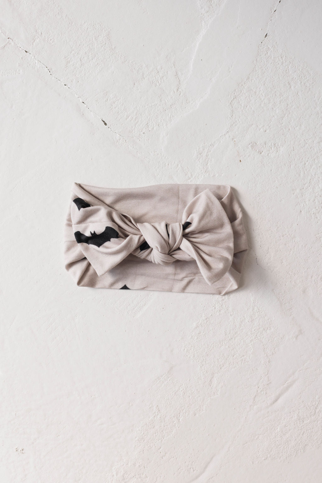 The "Bamboo Head Wrap | It's Bats!" by forever french baby, a beige headband with a twisted knot design, lies flat on a lightly textured, off-white surface. Crafted from bamboo material, the fabric features a subtle black pattern and is an adjustable head wrap perfect for babies and toddlers.