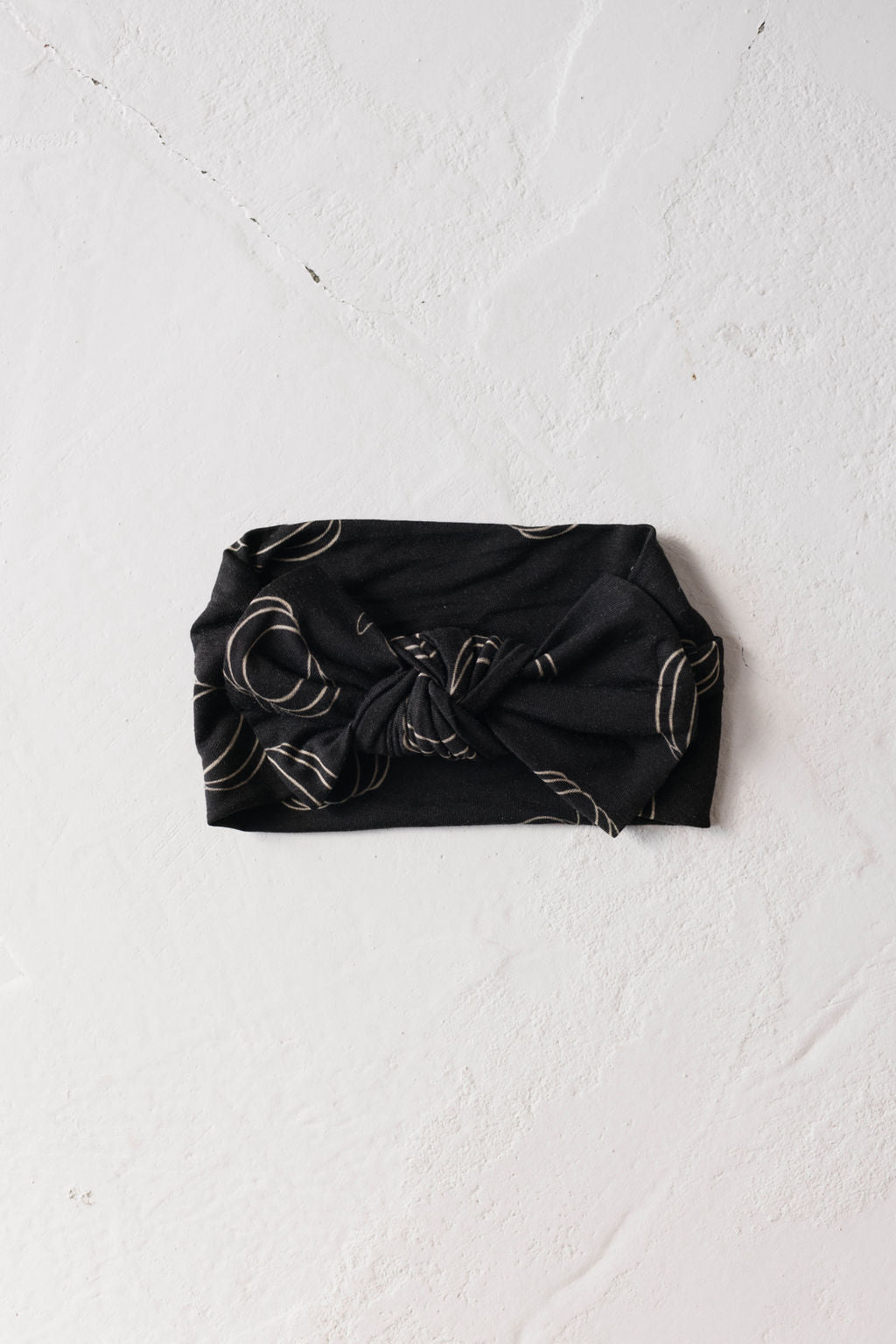 A Bamboo Head Wrap in the Midnight Pumpkin style from forever french baby, featuring a front knot and a subtle circular pattern, lies flat on a white, textured surface. This chic accessory is perfect for stylish babies and toddlers.