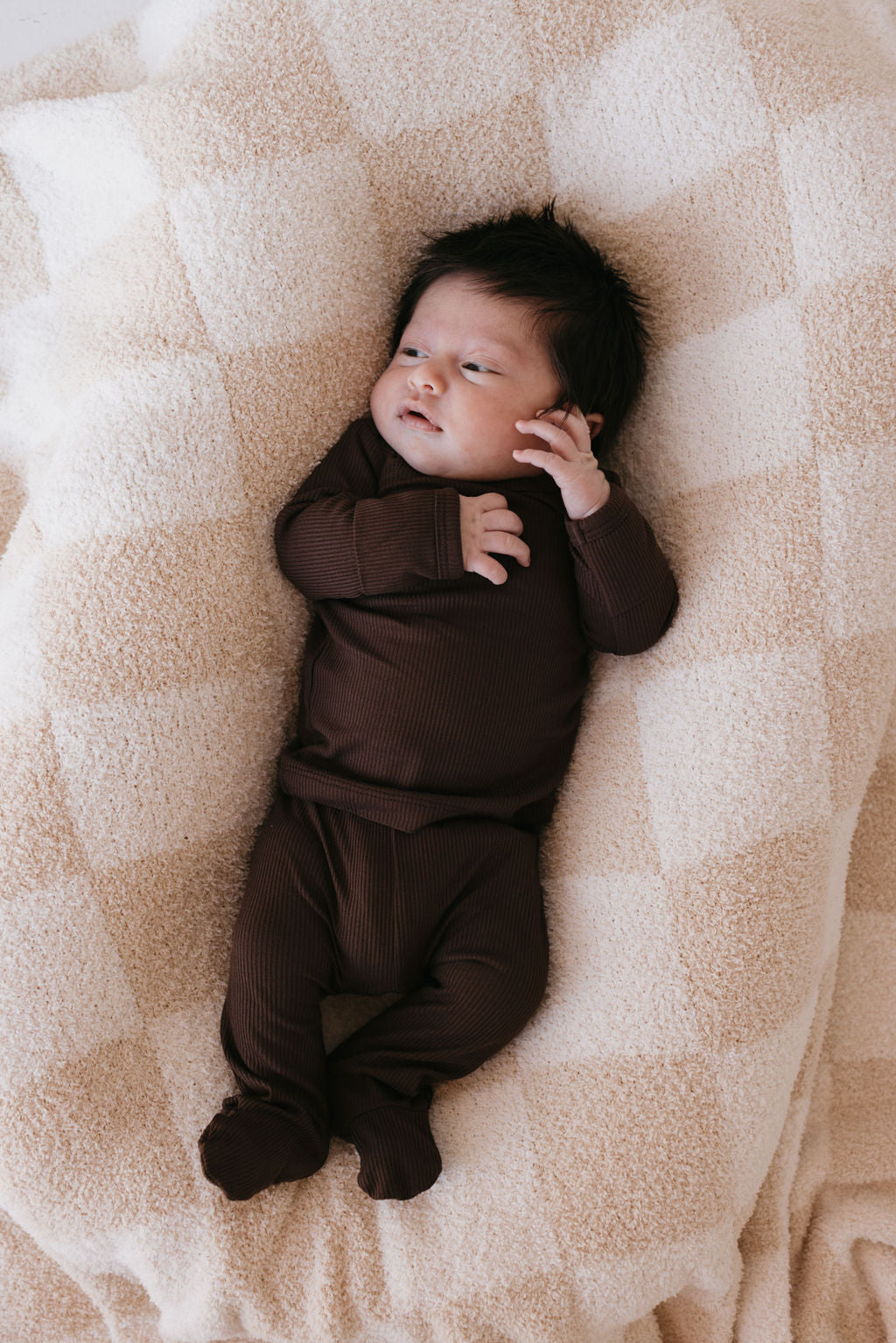 A baby in a Sleepy Time Set | Coffee Bean by forever french baby lies on a soft, checkered beige blanket. The infant has dark hair and a curious expression, with one hand near the face. This hypo-allergenic ensemble is perfect for sensitive skin.