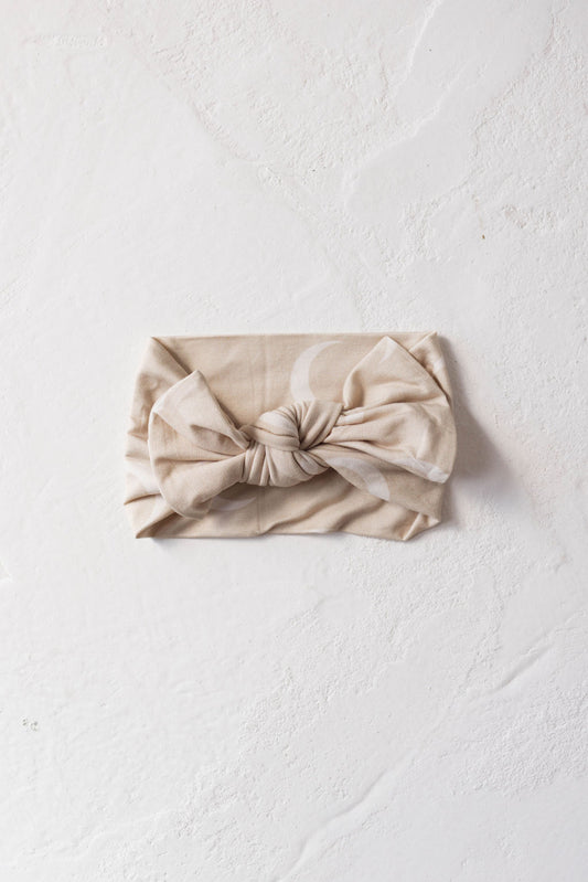 A beige, fabric headband with an interlocking knot design is laid flat against a textured white background. Perfect for your little one, this Forever French Baby Bamboo Head Wrap | Luna stylishly fits babies and toddlers.