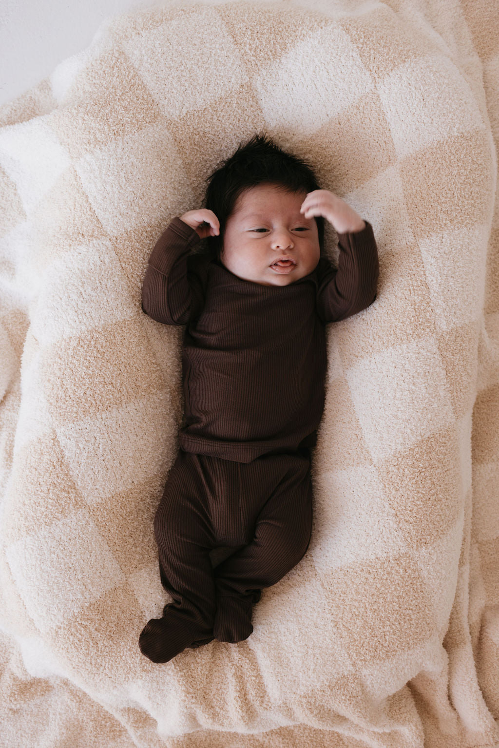 A baby dressed in the Sleepy Time Set | Coffee Bean from forever french baby lies on a soft, breathable checkered beige blanket, gazing upwards with its arms slightly raised.