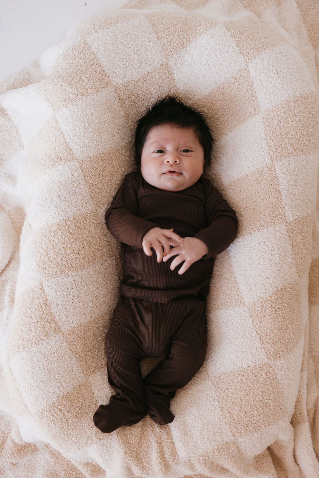 A baby dressed in the Sleepy Time Set in Coffee Bean from forever french baby lies on a soft, beige checkered blanket. With dark hair and clasped hands, the baby gazes upwards peacefully. The scene exudes serenity while highlighting the breathable, hypo-allergenic comfort of the outfit.