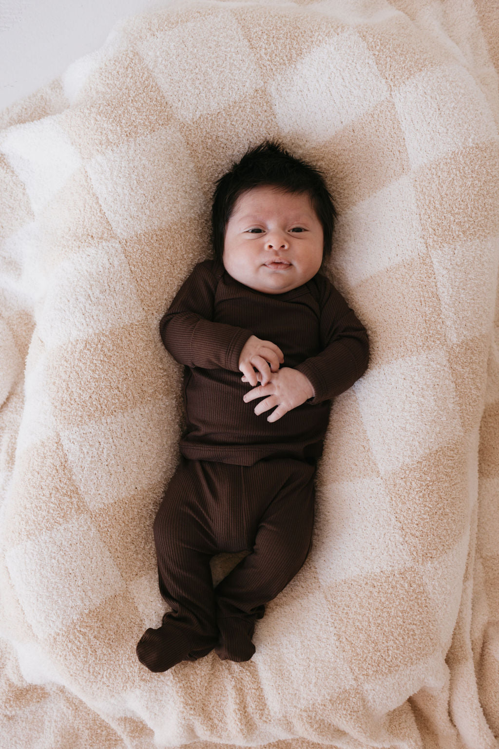 A baby in a Sleepy Time Set | Coffee Bean from forever french baby is lying on a cream-colored, checkered blanket. The baby has dark hair and a calm expression, with hands gently resting on the chest against the soft, hypoallergenic background.