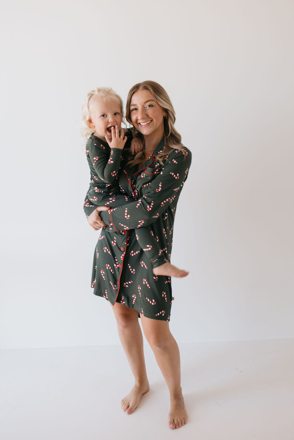 A woman and a child wearing matching pajamas from forever french baby's Bamboo Two Piece Pajamas | Candy Cane Lane collection exude joy. The woman, smiling with one hand on her hip, stands alongside the child who gazes curiously at the camera. Both are barefoot against a stark white background, enjoying their breathable sleepwear adorned with red and white candy cane patterns on dark green fabric.
