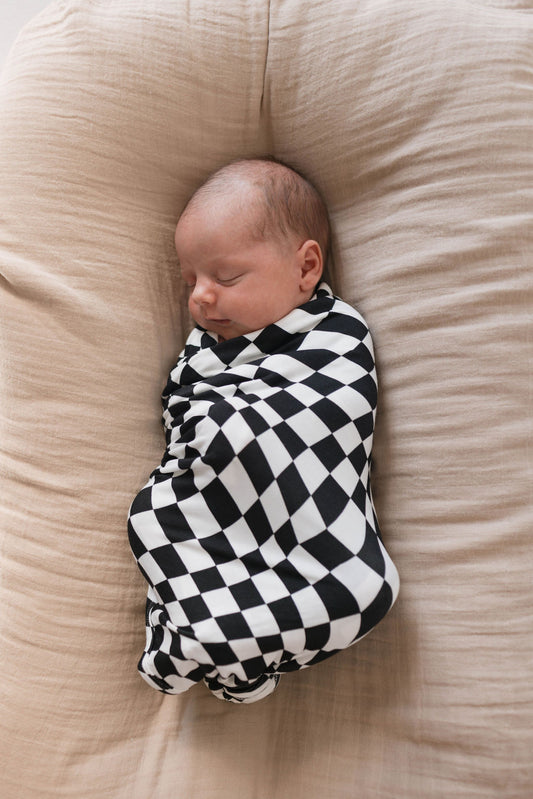 A newborn baby is peacefully sleeping while swaddled in a Black & White Wavy Checkerboard Bamboo Swaddle by forever french baby. The baby is lying on a soft, light beige cushion.