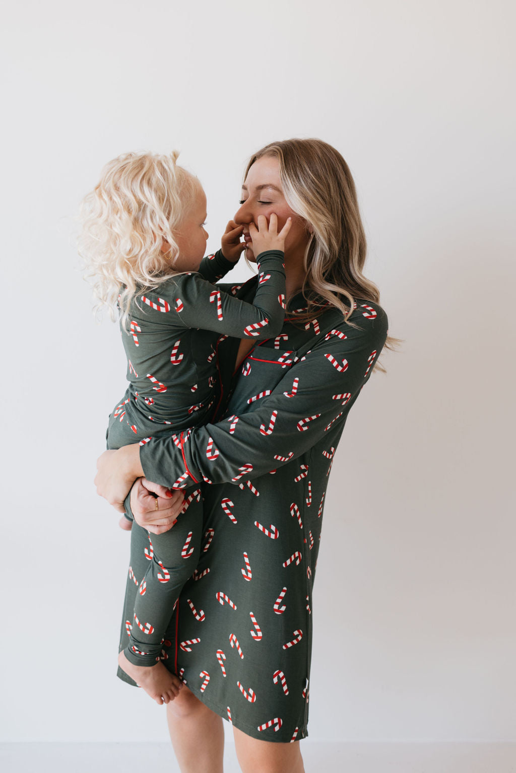 A woman cradles a child in her arms, both wearing matching green Bamboo Two Piece Pajamas from the Candy Cane Lane collection by forever french baby. The breathable sleepwear adds comfort as the child playfully touches the woman's face, their smiles bright against the plain white background.