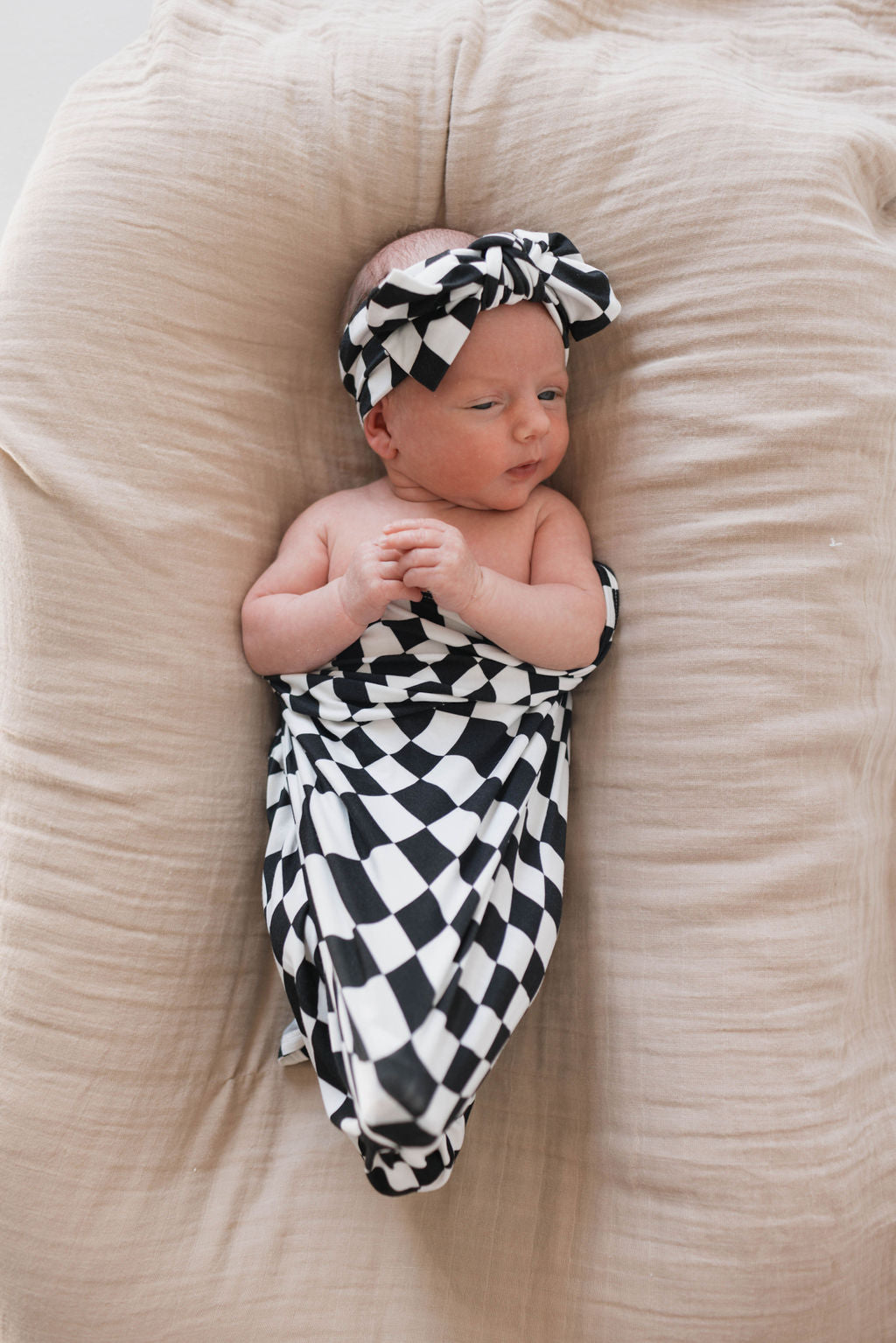 A newborn baby is lying on a beige cushion, wrapped in a hypo-allergenic forever french baby Bamboo Swaddle with a Black & White Wavy Checkerboard pattern that leaves their shoulders exposed. The baby is also wearing a matching black and white checkered headband with a bow. Their hands are clasped together.