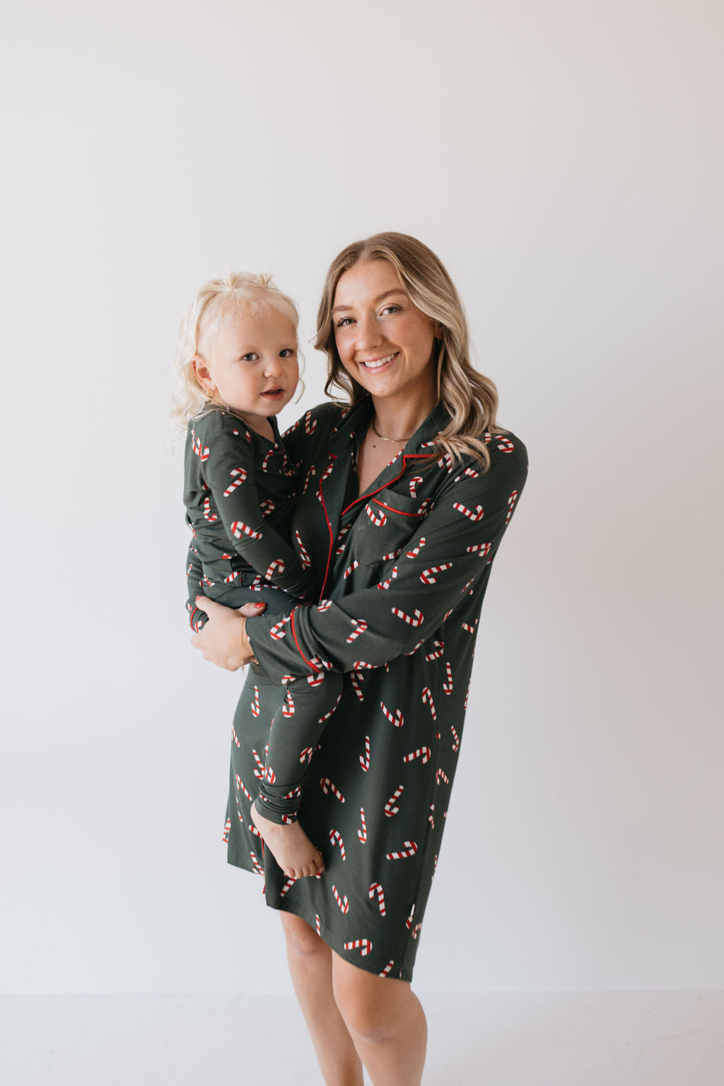 A woman is holding a young child, both dressed in matching Bamboo Two Piece Pajamas from forever french baby. The dark green pajamas feature festive red and white candy cane patterns, and they are smiling against a plain light background, relishing the comfort of their hypo-allergenic attire from the Candy Cane Lane collection.