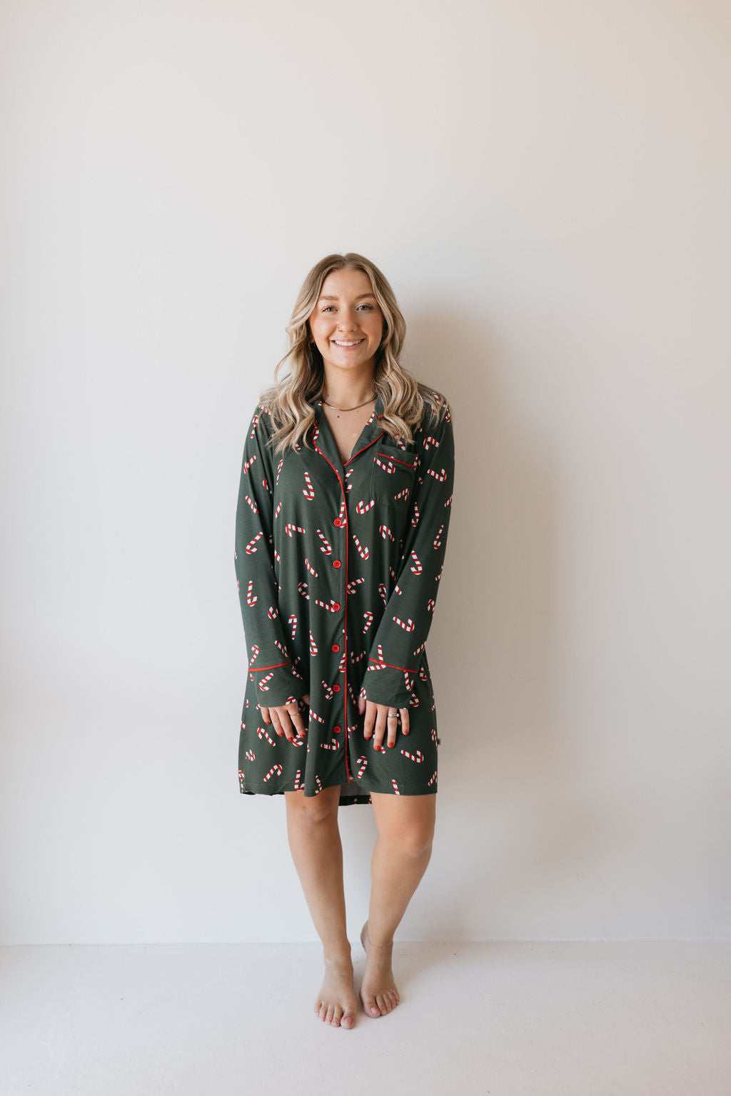 A woman stands smiling against a plain background, barefoot and adorned in the Women's Bamboo Sleeping Dress from forever french baby. The eco-friendly gown features a charming pattern of small red and white candy canes, with her long, wavy hair cascading gracefully over her shoulders.