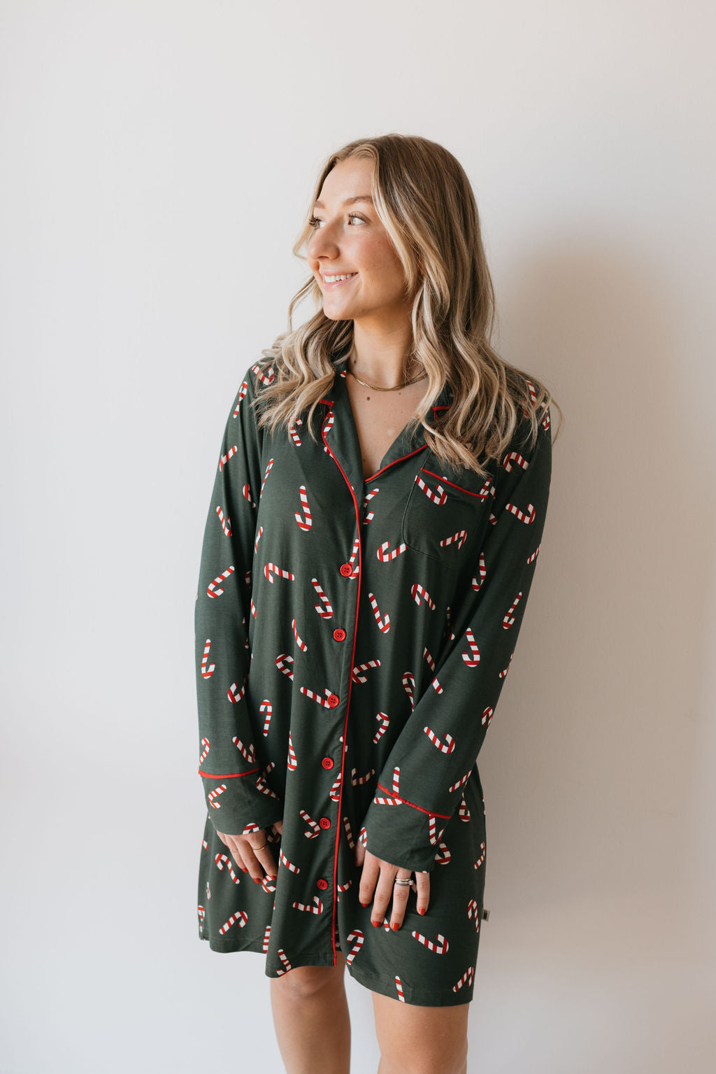 A woman in a Women's Bamboo Sleeping Dress, featuring the Candy Cane Lane design by forever french baby, stands against a plain background, smiling and gazing to the side.